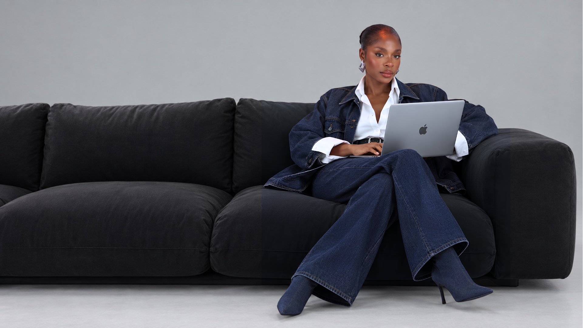 Abigail in a denim suit and white shirt, working on a laptop while seated on a black couch
